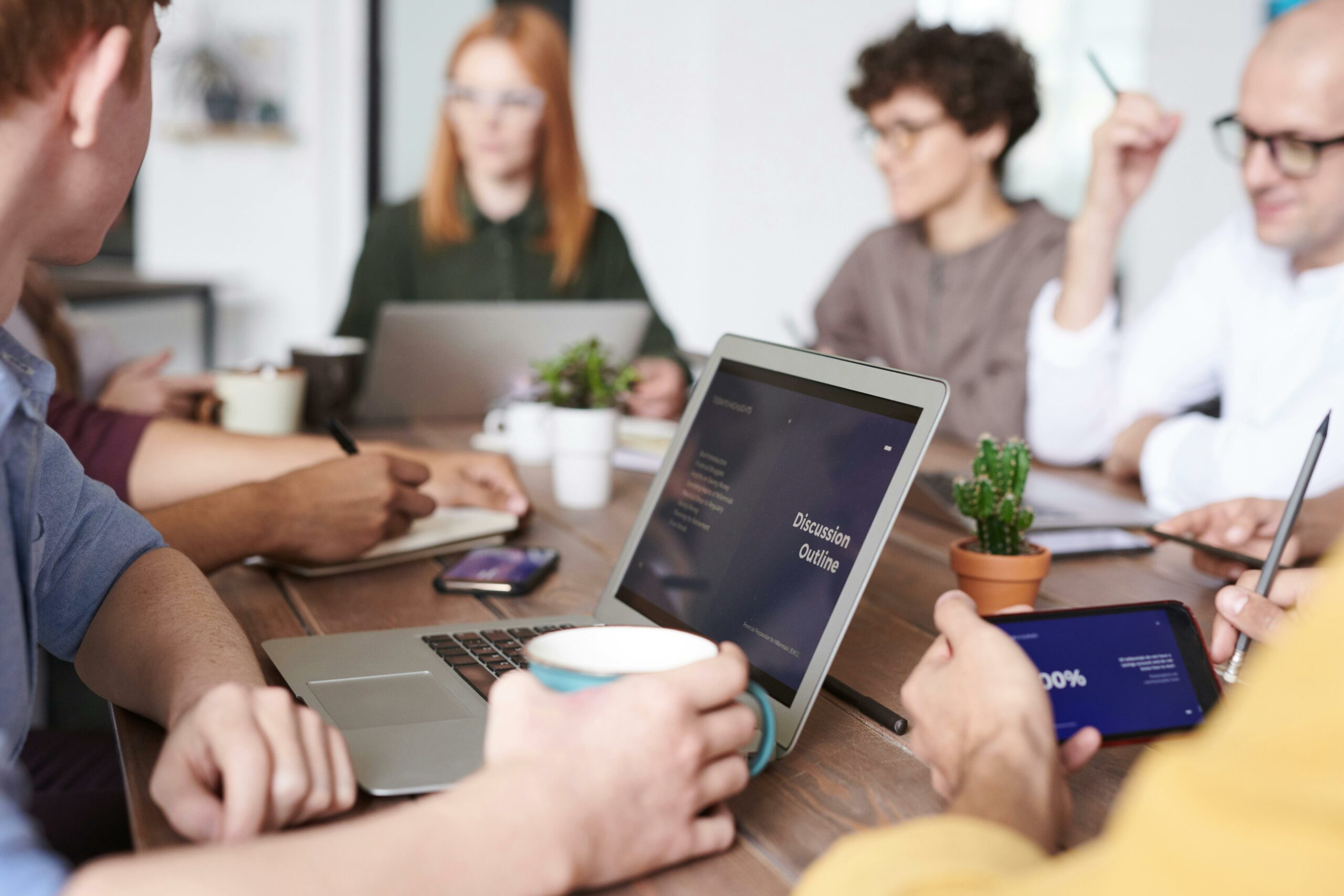 Group of professionals collaborating at a coworking space table with laptops, coffee mugs, and a discussion outline on screen, representing community and innovation.